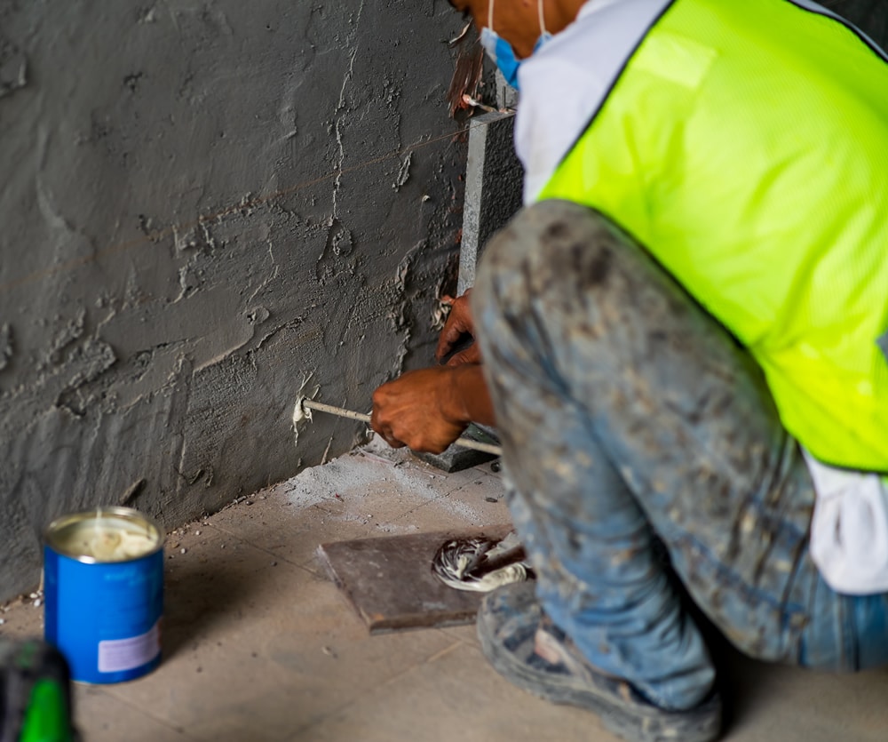 Worker applying damp-proofing treatment to a wall, wearing a safety vest and mask, with tools and materials visible on the floor, illustrating damp proofing process.