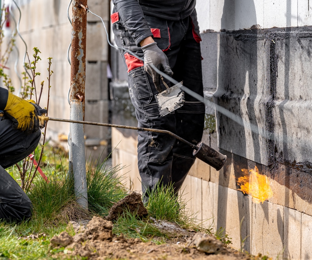 Workers applying damp proofing treatment to a building's foundation using a torch and trowel, highlighting the damp proofing process essential for moisture protection.
