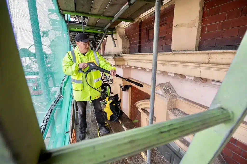 Worker in high-visibility jacket using a pressure washer for brick cleaning on scaffolding, showcasing restoration techniques by Environ Restoration Services in London.