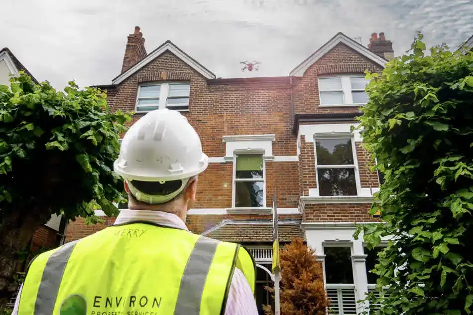 Construction worker in safety gear observing a drone surveying a building roof, highlighting advanced drone technology for efficient roof inspections by Environ Restoration Services.