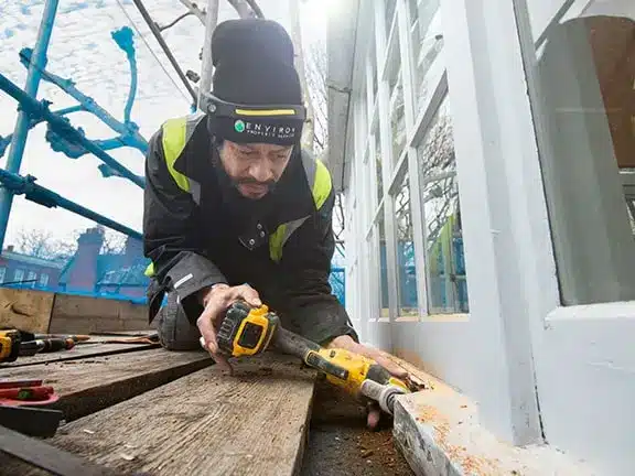 Worker repairing timber window with power tools, focused on restoration techniques, scaffolding in background, highlighting expertise in period property maintenance.