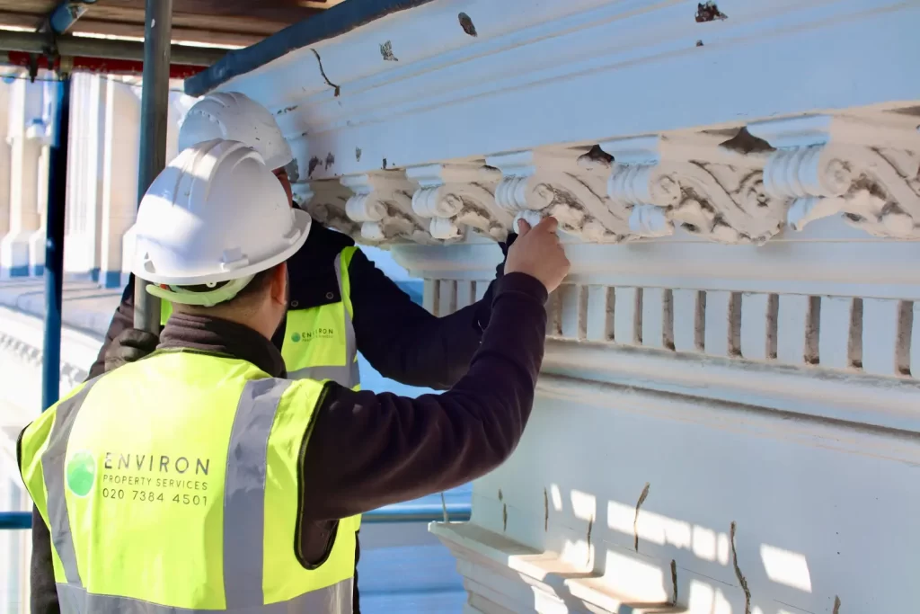 Two professionals from Environ Restoration Services cleaning and restoring decorative stone masonry, wearing safety helmets and high-visibility vests, on scaffolding in a historical building context.
