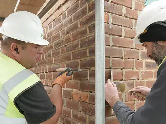 Two workers in hard hats and safety gear repointing a brick wall, demonstrating techniques for mortar application and repair.