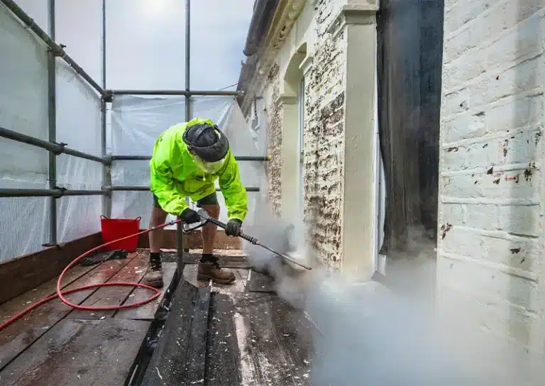 Professional facade cleaning in progress, worker using high-pressure cleaning equipment to remove dirt and grime from building exterior, scaffolding and protective sheeting visible.