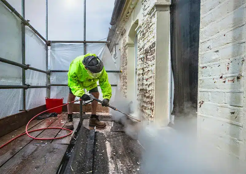Worker in high-visibility jacket using pressure washer on historic building facade, surrounded by scaffolding and protective sheeting, illustrating restoration techniques for historic property surveys in London.