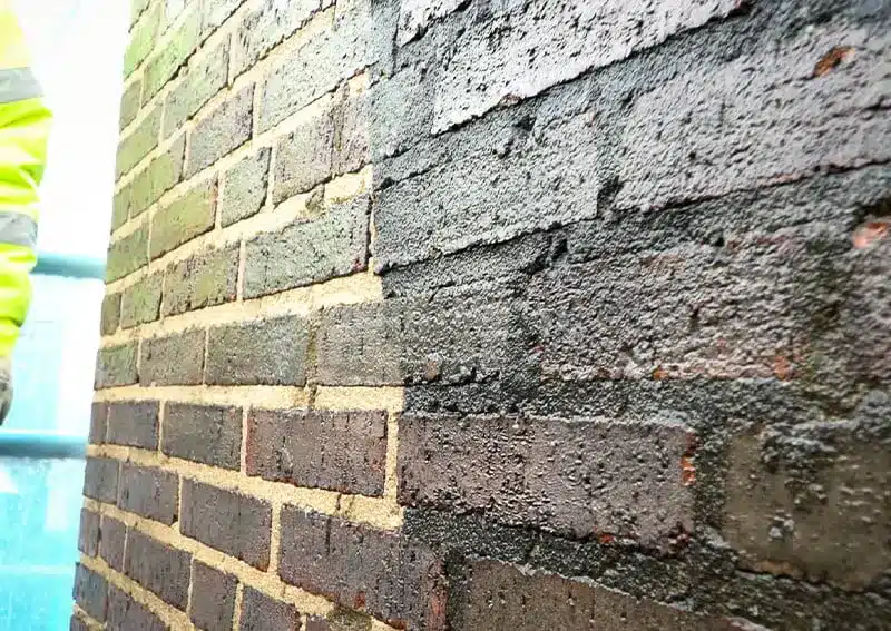 Brick wall showing contrasting sections of paint removal, highlighting restored brick texture and colour, with a worker in a high-visibility jacket partially visible.