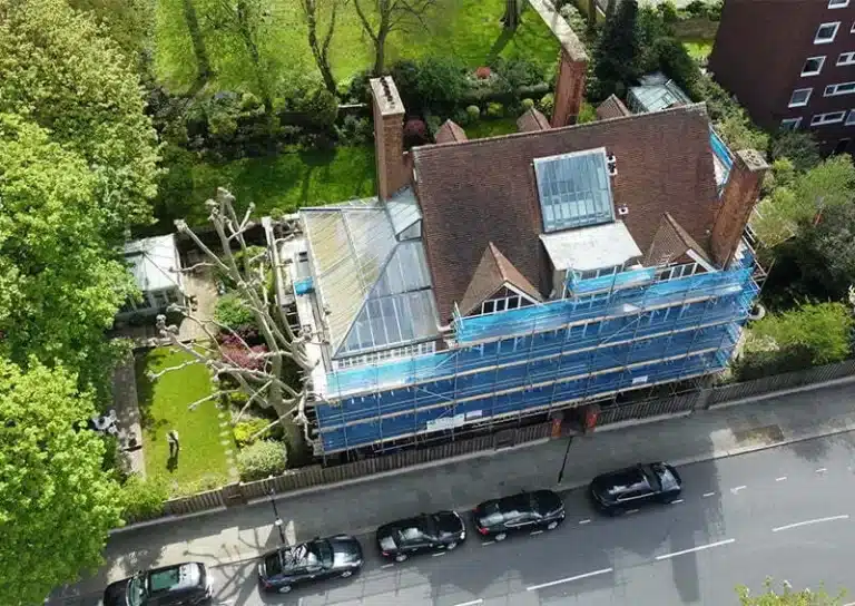 Aerial view of a residential property under renovation, featuring scaffolding and protective coverings, surrounded by green gardens and parked cars, illustrating the importance of professional damp survey services in London.