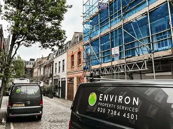 Environ Property Services vans parked on a cobblestone street, with scaffolding and restoration work visible on a building, reflecting expertise in Section 20 works for communal properties in London.