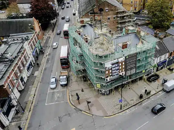 Building undergoing restoration with scaffolding and protective covering, located at a busy intersection in London, showcasing ongoing Section 20 works for communal properties.