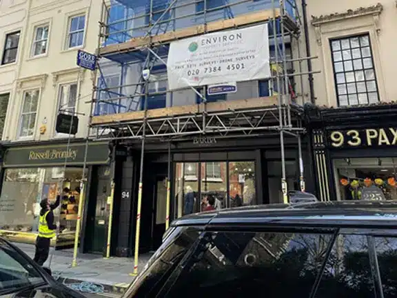 Scaffolded building with Environ Restoration Services banner, showcasing ongoing Section 20 works in London, including construction staff and nearby shops.