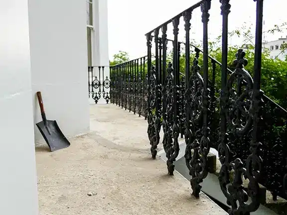 Shovel resting against a decorative wrought iron railing on a stone surface, suggesting preparation for stone wall restoration work.