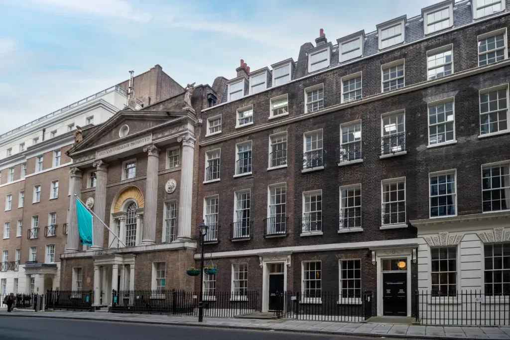 Historic London buildings with classical architecture, showcasing a grand entrance with columns and a blue flag, surrounded by modern structures, reflecting the city's blend of old and new.