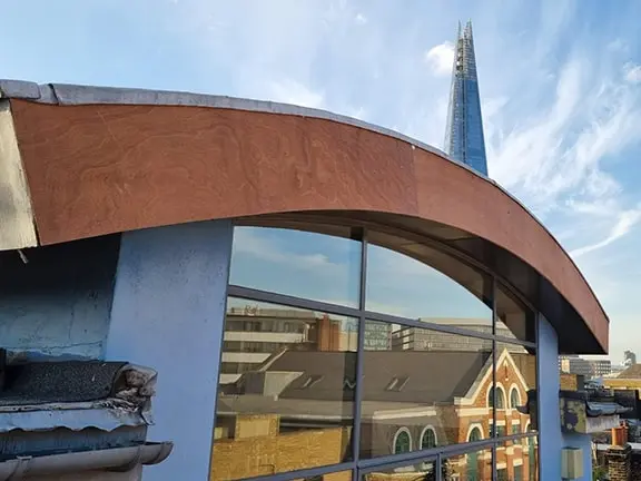 View of a listed building restoration project featuring a modern curved roofline with reflective windows, overlooking historic architecture and The Shard in London, illustrating the blend of traditional and contemporary design elements.