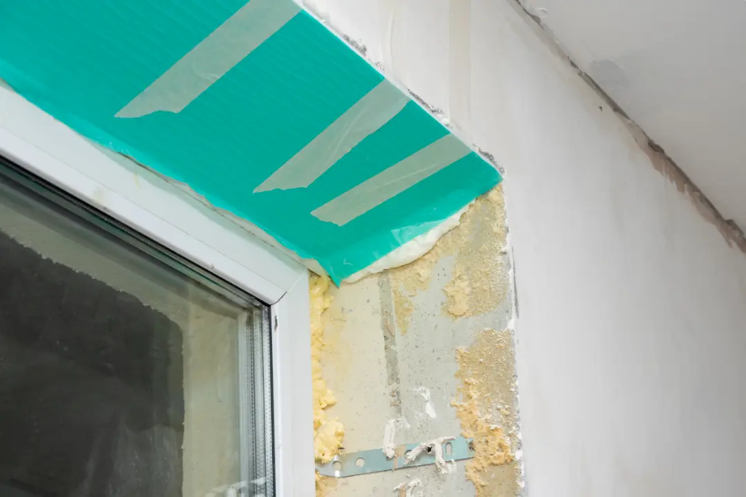 Close-up of a window corner showing exposed wall with foam insulation, masking tape, and unfinished plaster, illustrating potential restoration issues in a building.